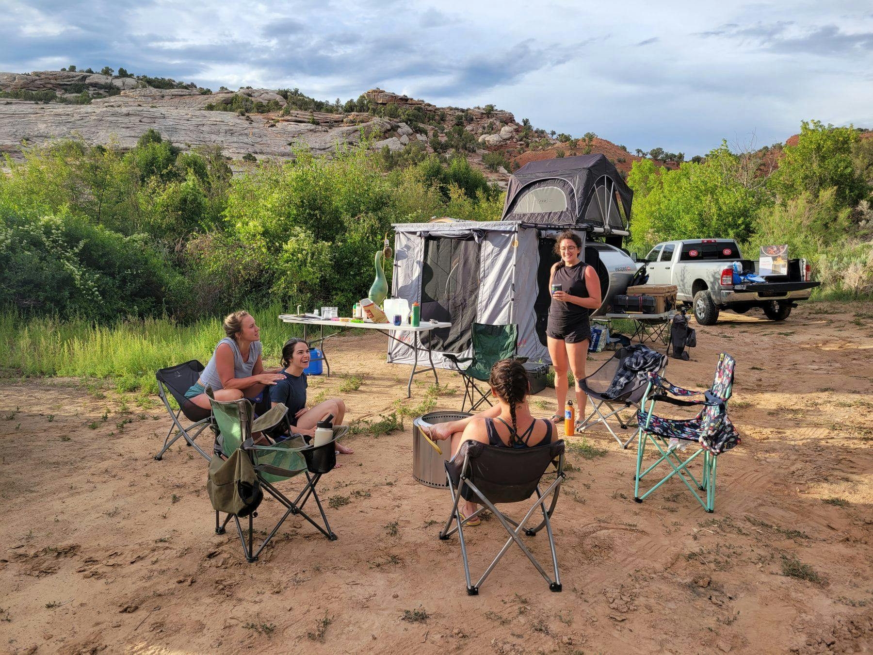 A group of women sitting around a fire in front of a teardrop camper.