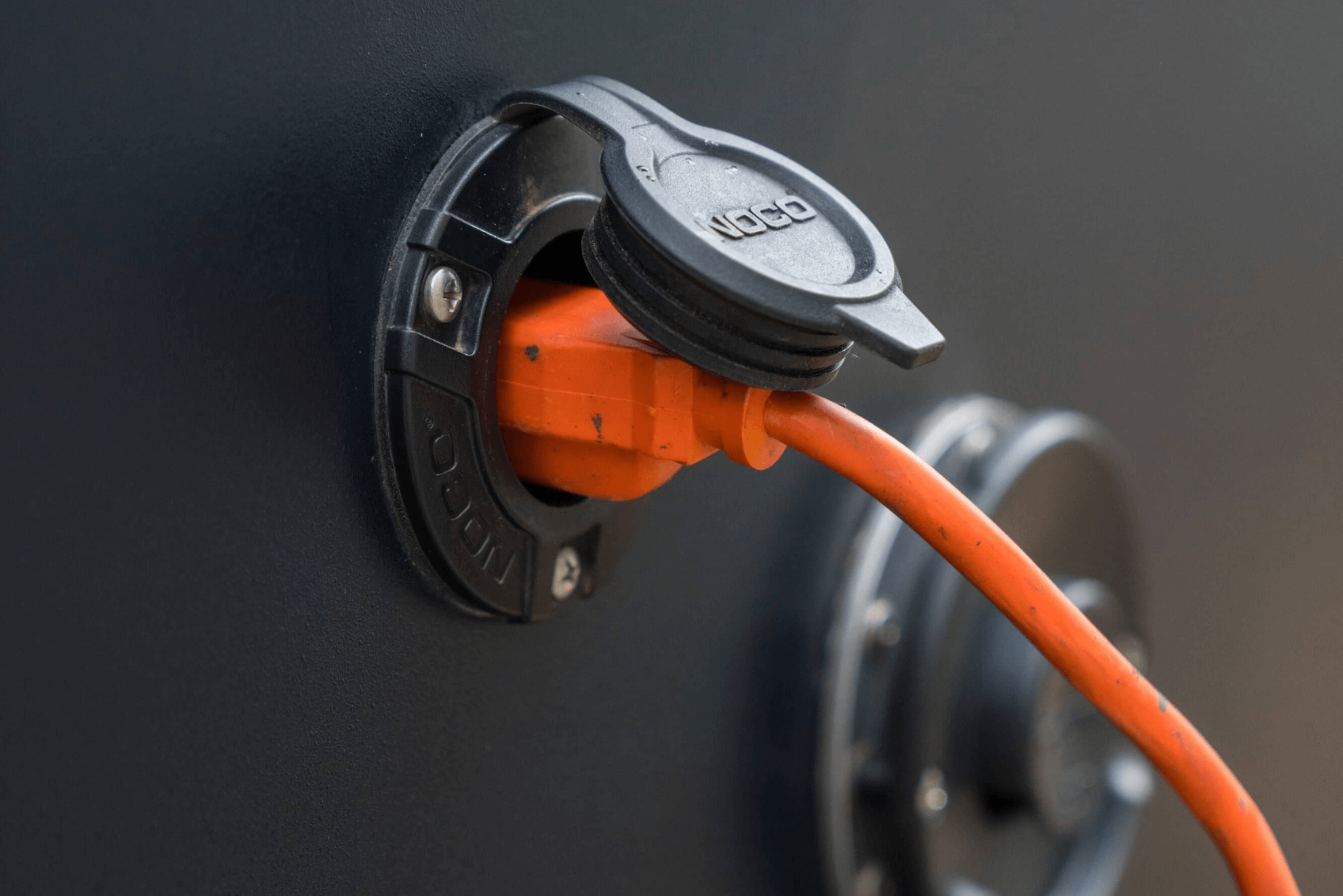 An orange extension cord being plugged into a NOCO shore power port on the side of a teardrop camper.