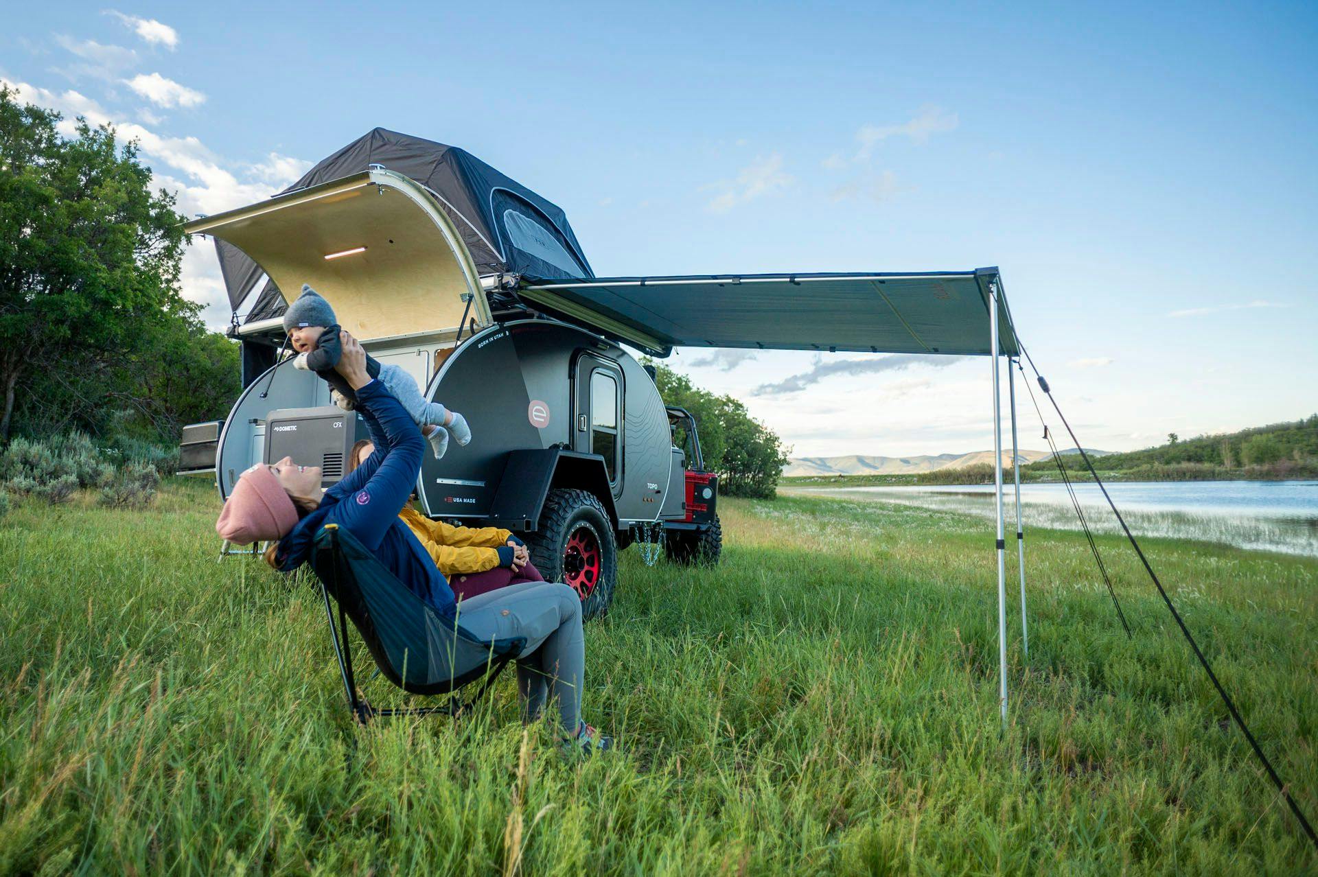 Two women and a small child enjoying their campsite next to a teardrop camper.