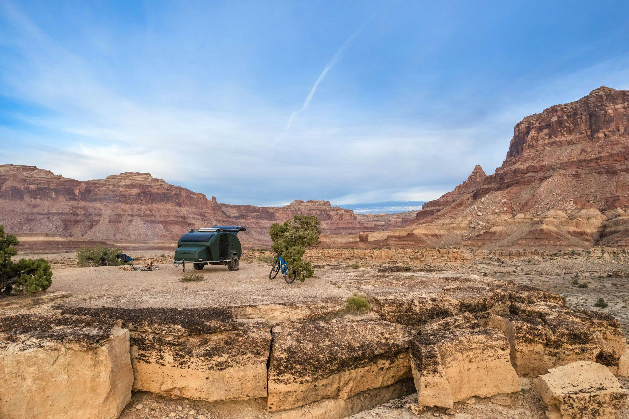 A green teardrop trailer parked on the ledge in the desert. A blue mountain bike resting closely beside it.