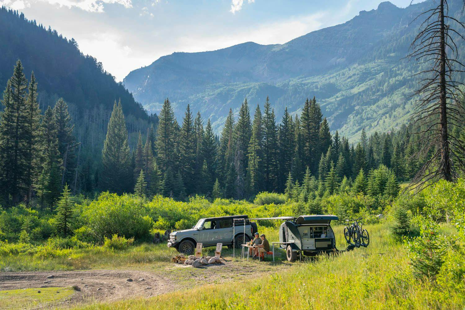 A teardrop camper set up in a beautiful campsite with green grass, pine trees, and mountain all around.