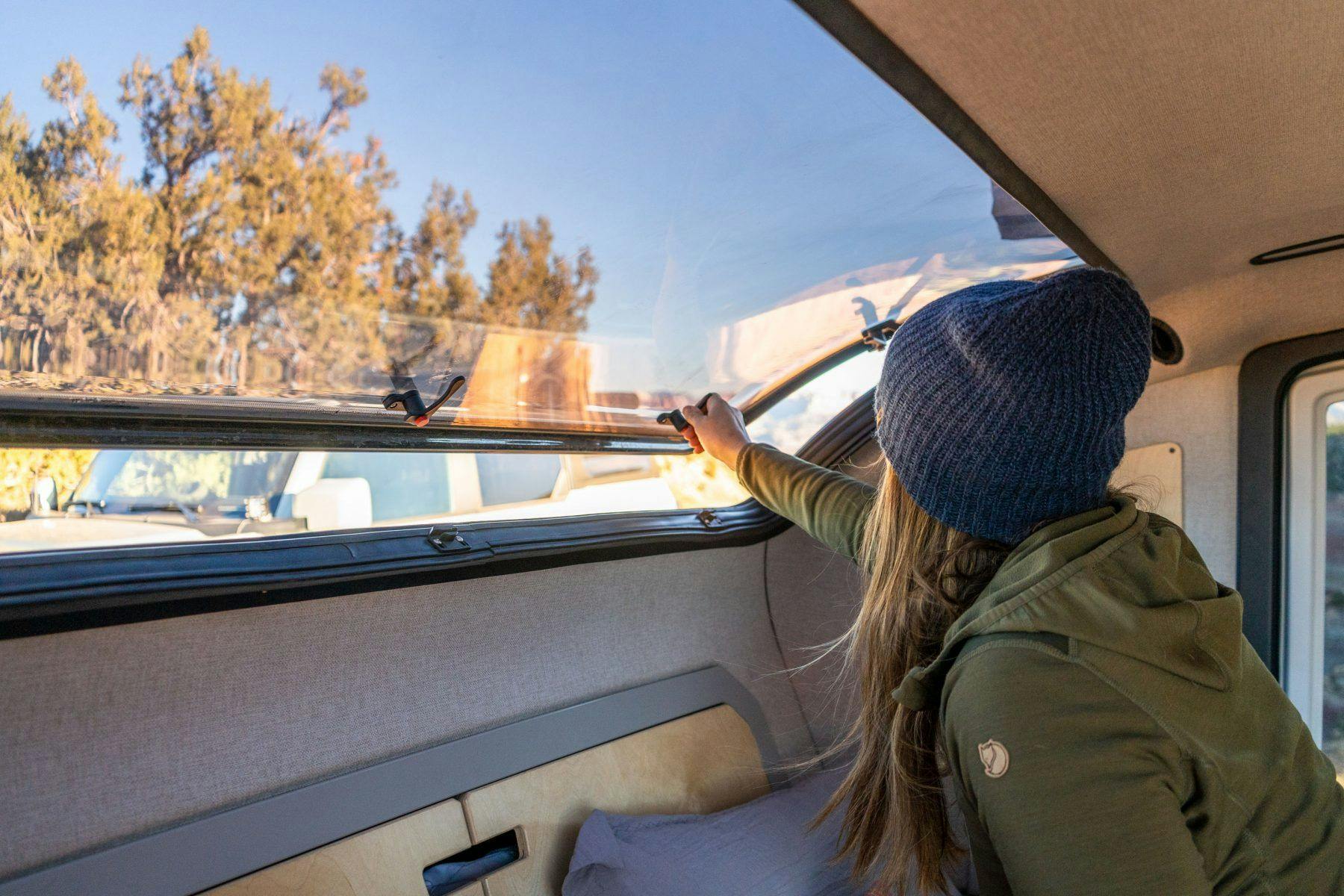 A woman wearing a green jacket and blue beanie opening the the stargazer of a teardrop camper.