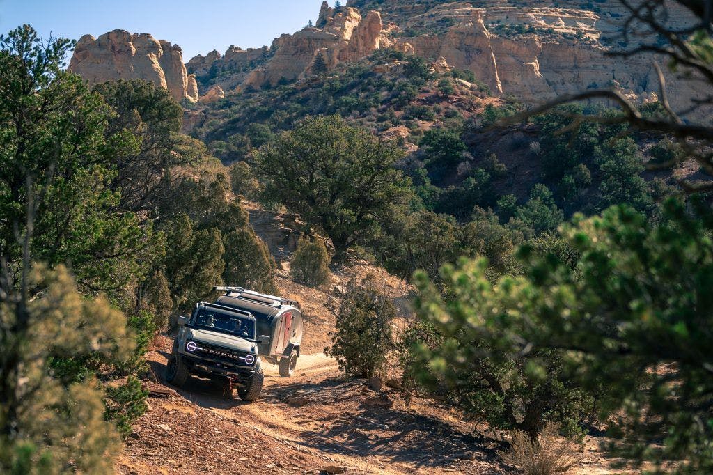 A navy blue teardrop camper being towed through desert landscape by a Ford Bronco.