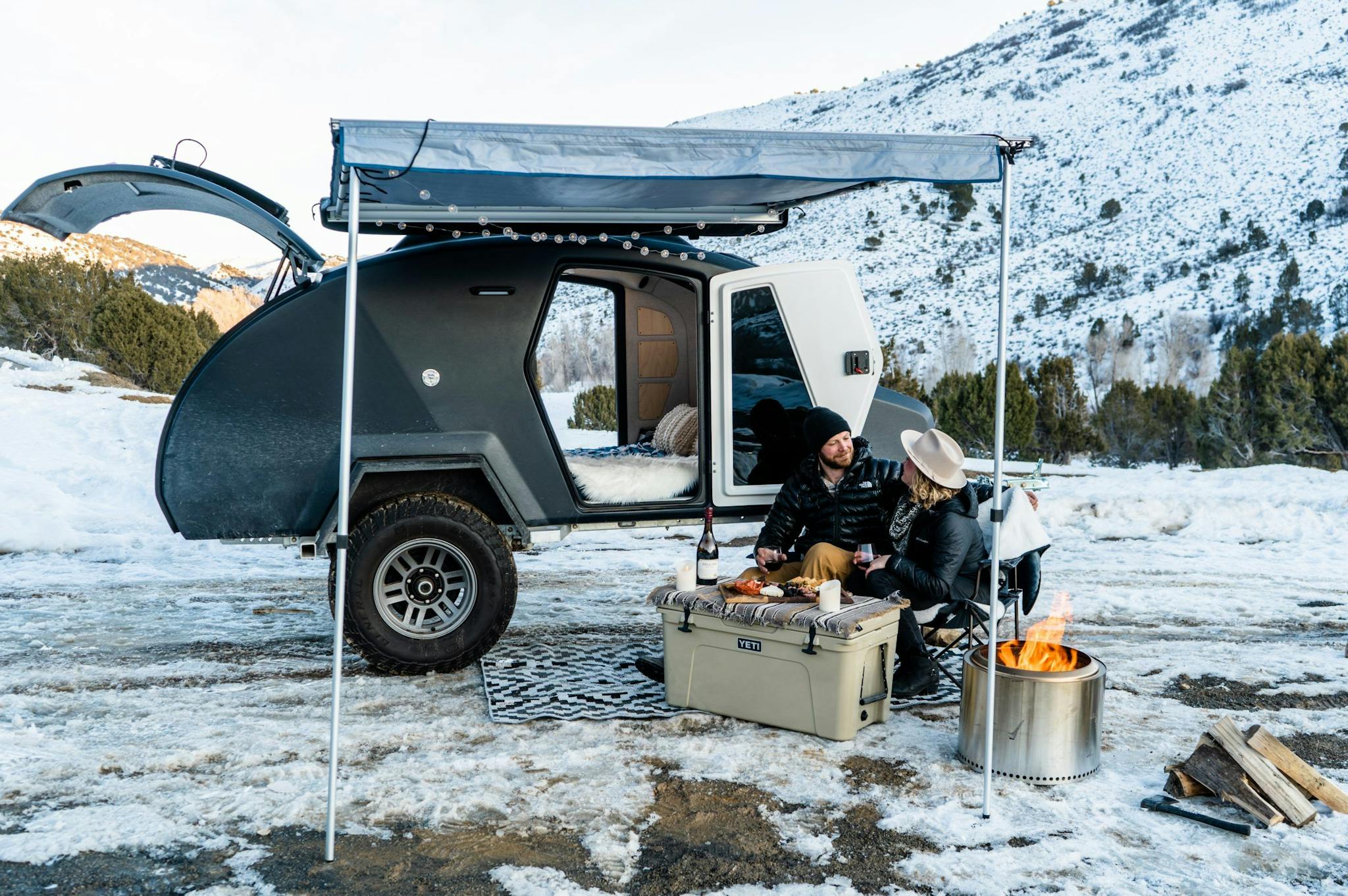 A couple sitting outside their navy teardrop camper in winter landscape.