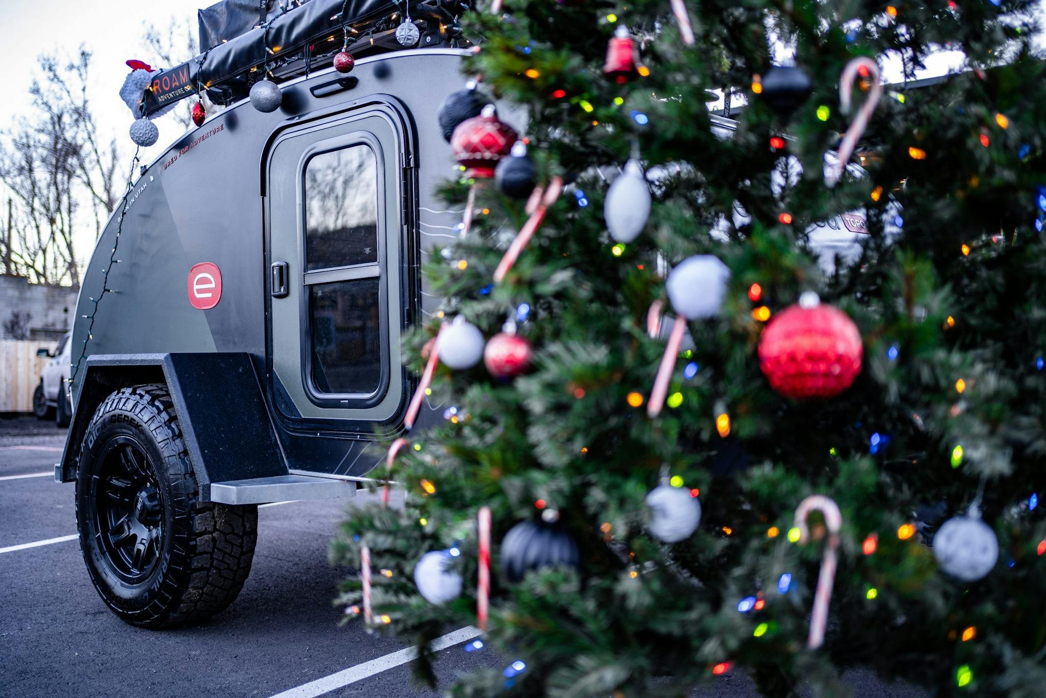 A beautiful christmas tree decorated with silver & red ornamets with a teardrop trailer tucked behind the tree.