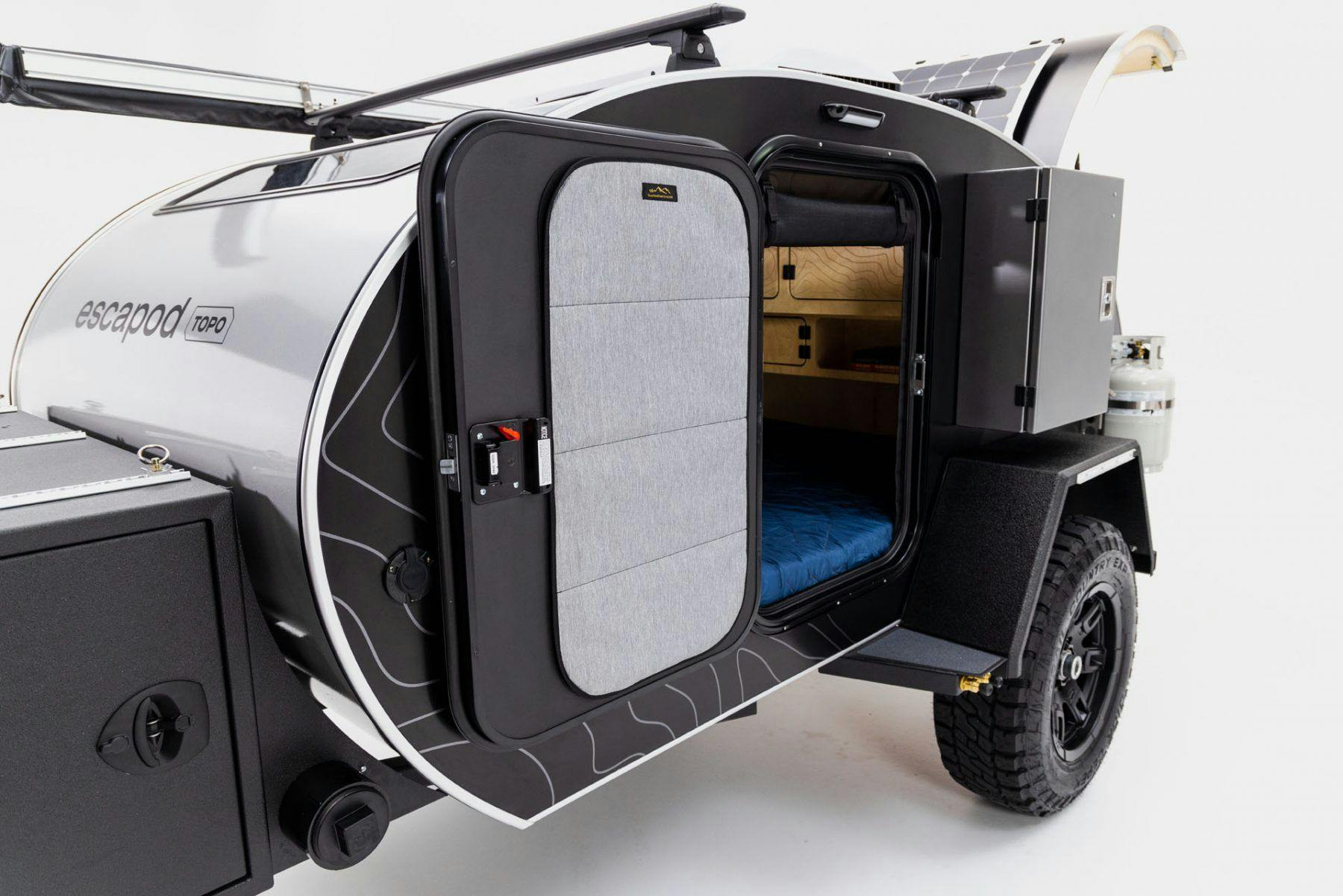 An Escapod Original TOPO teardrop camper in a studio against a white backdrop. A solar panel and spare tire accent the trailer.