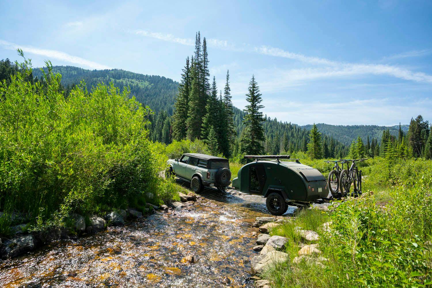 A teardrop trailer with bikes on board, being towed across a water crossing by a Ford Bronco.