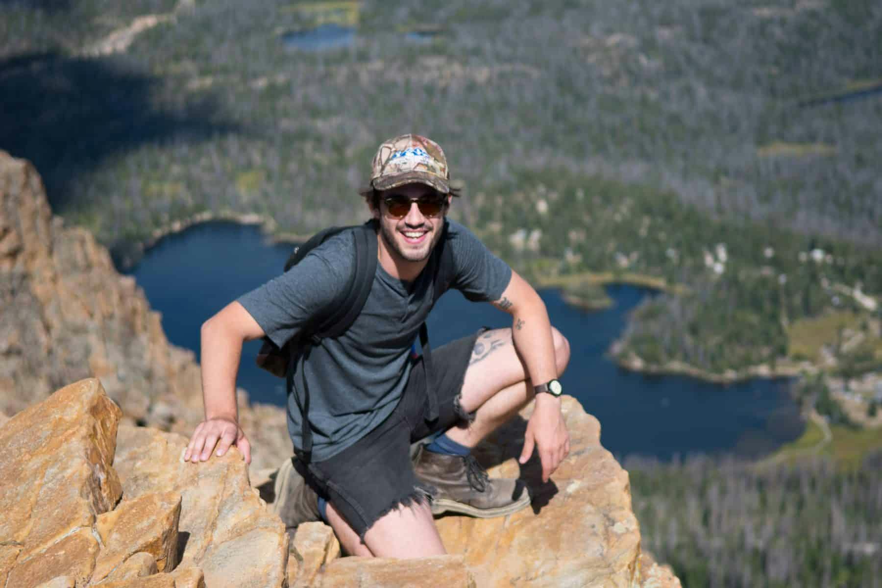 A man at the peak of a mountain with a display of a lake behind him.