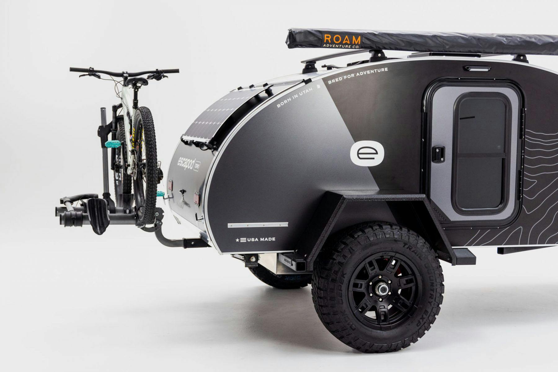 An Escapod Original TOPO teardrop camper in a studio against a white backdrop. A solar panel and spare tire accent the trailer.