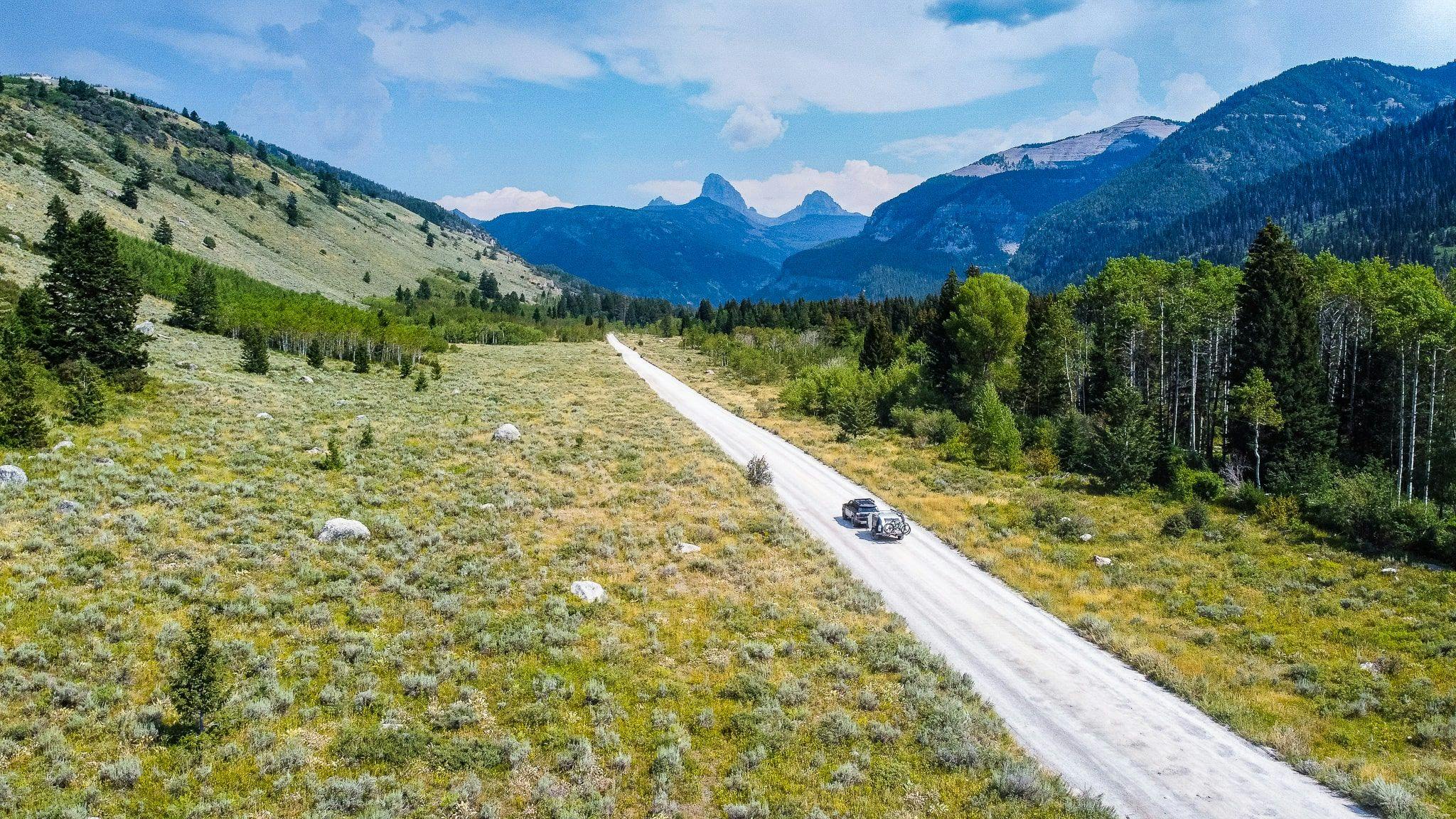 A teardrop trailer being towed on a backroad through an area with sagebrush, trees, and mountains.