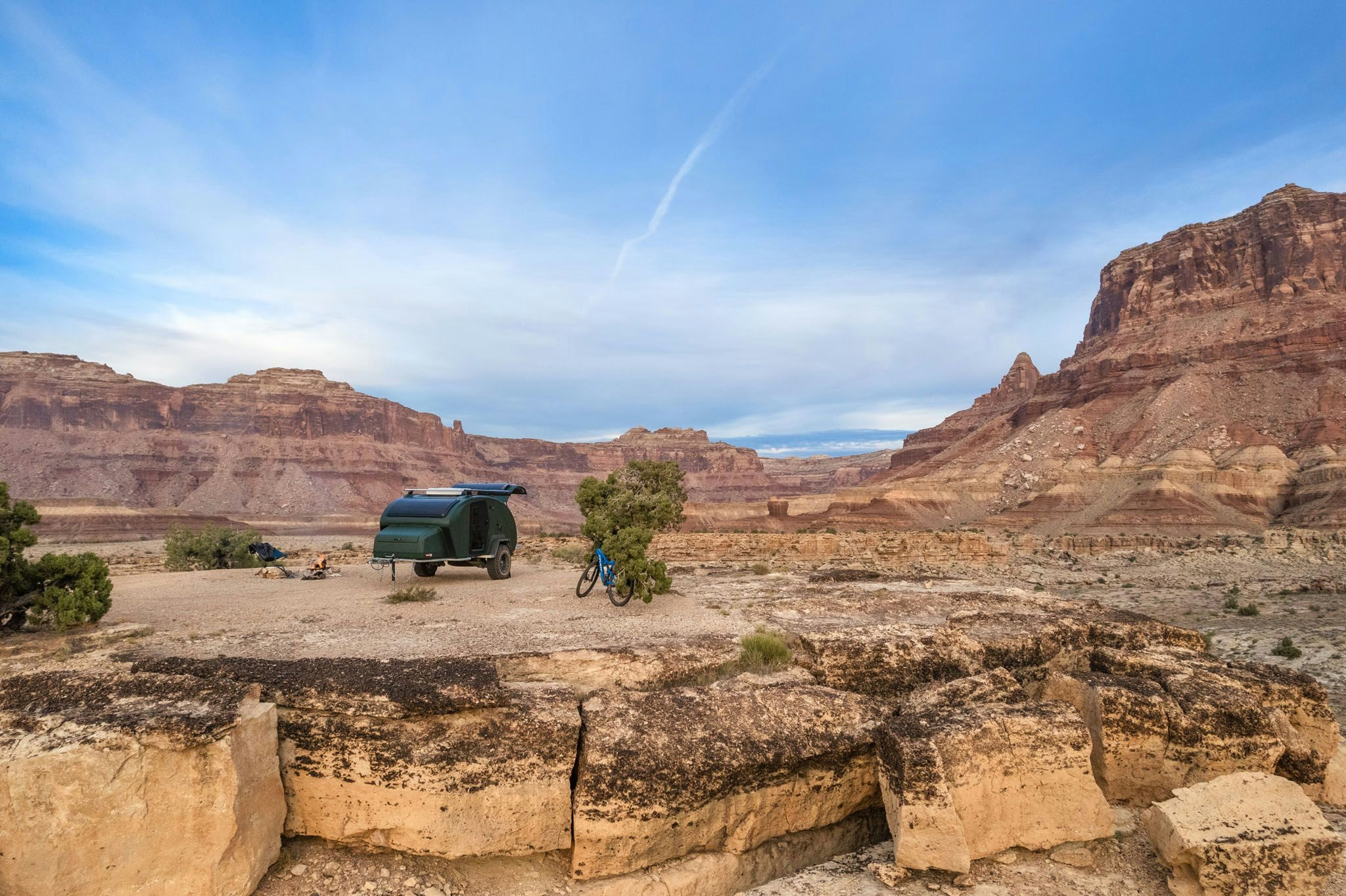 A green teardrop trailer parked on the ledge in the desert. A blue mountain bike resting closely beside it.