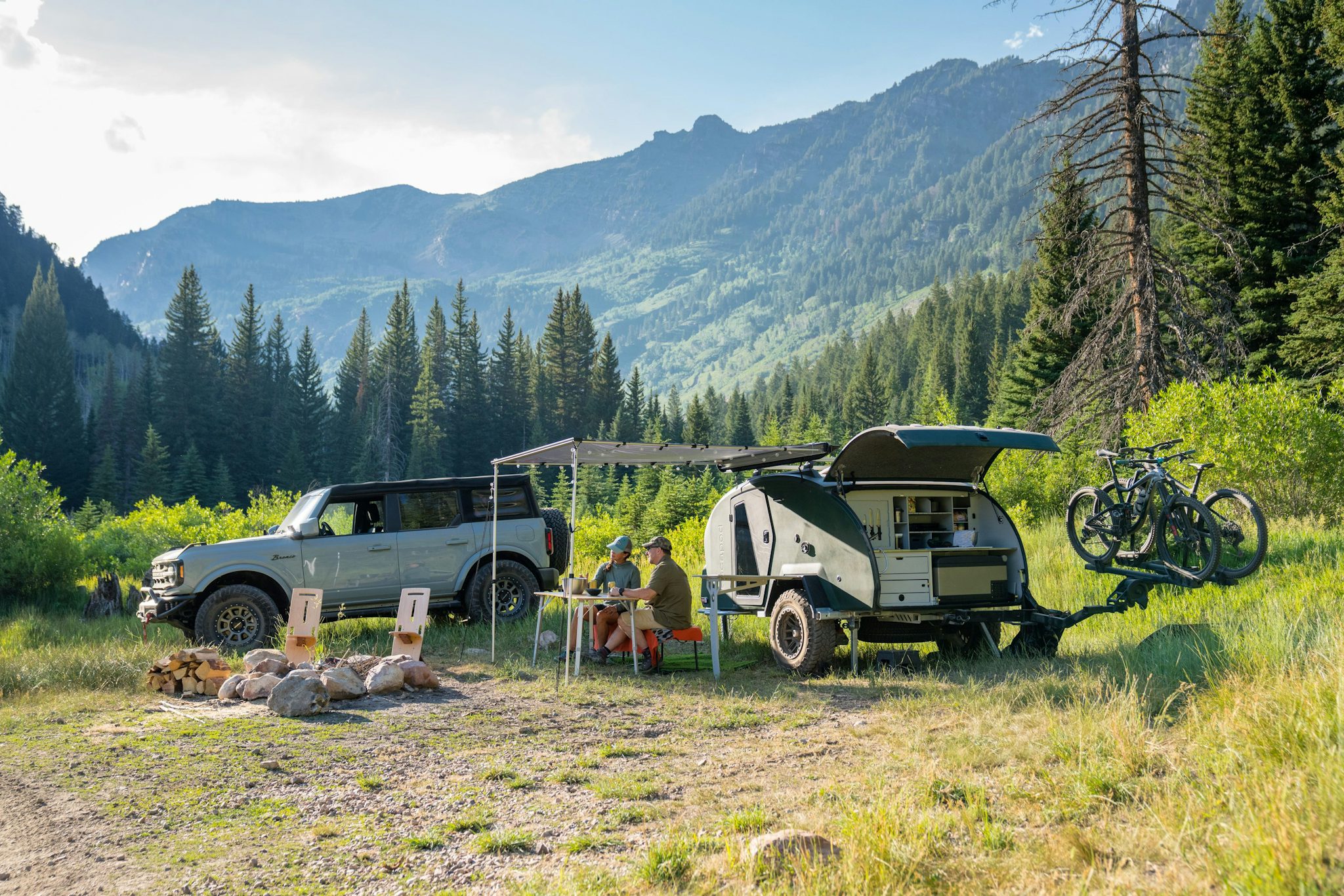 A teardrop trailer set up in a beautiful green field, surrounded by trees, mountains, and blue skies.